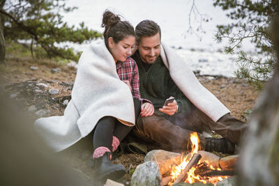 Couple using mobile phone while wrapped in blanket by fire pit at campsite