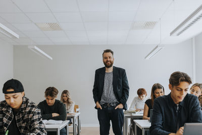 Portrait of smiling professor standing with hands in pockets amidst students