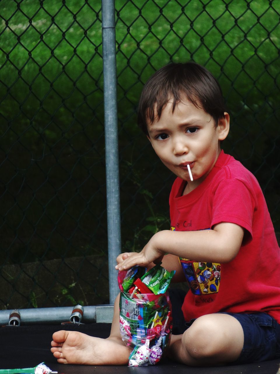 HAPPY BOY HOLDING CHAINLINK FENCE AGAINST BLURRED GREEN LANDSCAPE