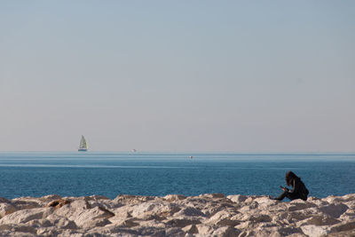 Woman sitting on rocks by sea against clear sky