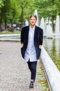 Full length portrait of young woman in blazer by pond on footpath at park