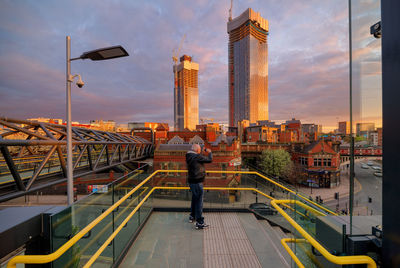 Man standing by railing in city against sky during sunset