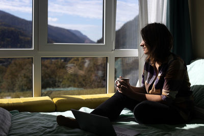 Young woman using phone while sitting on window