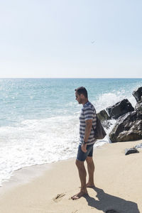 Side view of man looking at sea while standing by rocks at beach against clear sky
