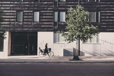 Side view of mature woman walking with bicycle on sidewalk against building in city
