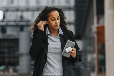 Young businesswoman wearing wireless in-ear headphones while holding smart phone