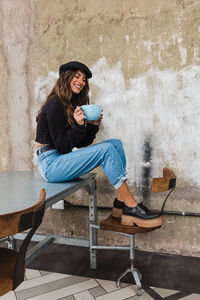 Young woman drinking coffee while sitting on chair against wall