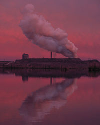 Smoke emitting from factory by lake against sky