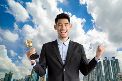 Portrait of cheerful businessman holding trophy while standing against sky