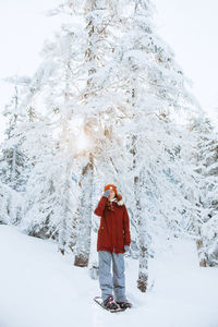 Full body woman in outerwear with snowshoes covering eye while standing near snowy spruces on cold winter day in valley of the ghosts in monts valin national park in quebec, canada