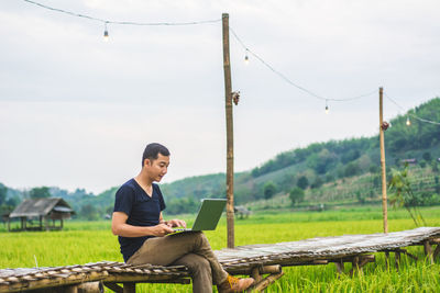 Man using laptop while sitting on boardwalk at rice paddy