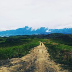 Dirt road amidst grassy field leading towards mountains against cloudy sky