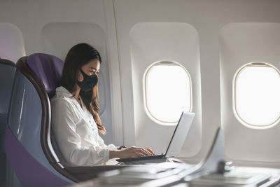 Smiling businesswoman wearing mask working on laptop while sitting in airplane