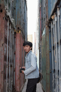 Portrait of man holding camera standing on alley amidst cargo containers