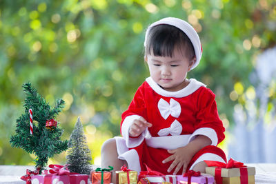 Full length of cute baby girl wearing santa costume while sitting with christmas decorations on table against trees