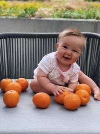 A baby girl with blond hair sitting on a table on a terrace with big oranges around her 