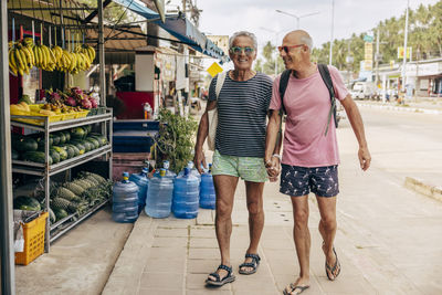 Smiling elderly gay couple holding hands while walking near supermarket at street
