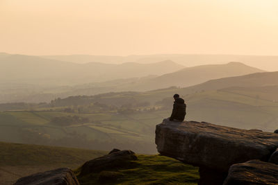 Man sitting on cliff against sky during sunset