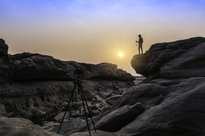 Man holding camera while standing on rock against sky during sunset