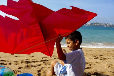 Boy playing with red kite at beach during sunny day