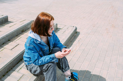 Young stylish woman walks on street with a skateboard and uses a smartphone