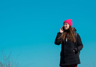 Young confident woman in pink hat smartphone in hand calling friend walking on blue sky background.