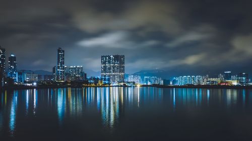 Reflection of blue illuminated buildings in river against sky at night