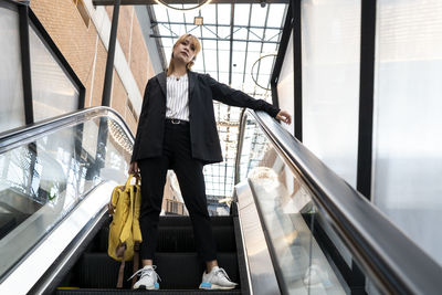 Low angle portrait of woman with backpack standing by railing on escalator