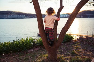 Rear view of boy standing on tree at lakeshore