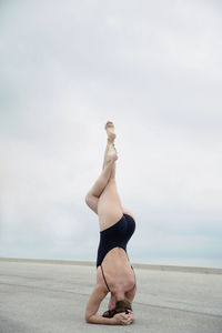 Rear view of flexible woman wearing one piece swimsuit while practicing headstand on road against sky