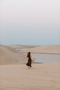 Woman in black dress strolling on sand towards water while spending time in desert in morning in doha, qatar