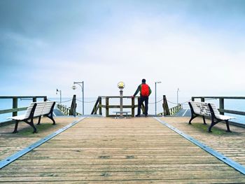 Tourist with red backpack on wooden pier above sea. man in trekking suit in harbor in rainy day