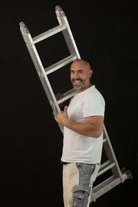 Portrait of smiling mature man holding ladder while standing against black background