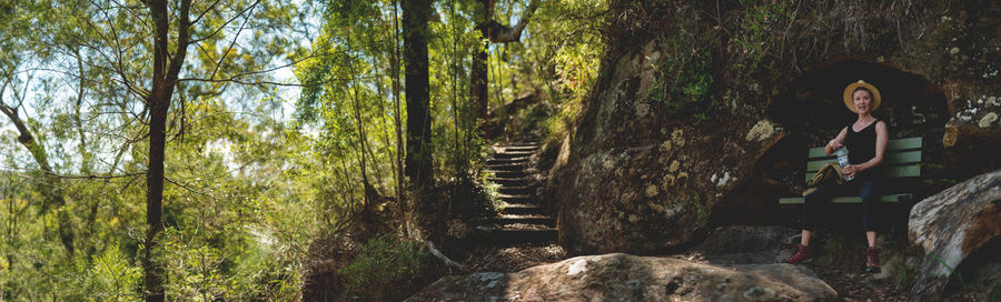 Panoramic view of woman sitting on bench against rock in forest