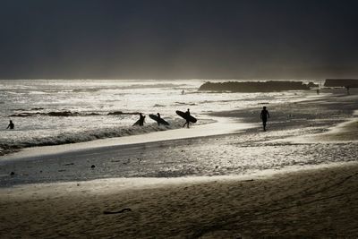 Silhouette surfers on shore at beach against sky during sunset