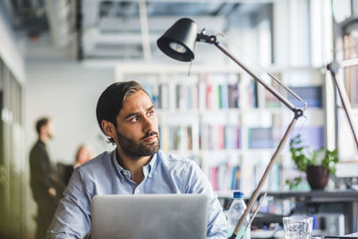 Thoughtful businessman sitting with laptop at desk in creative office