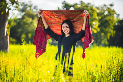 Portrait of young woman holding shawl while standing amidst grassy field