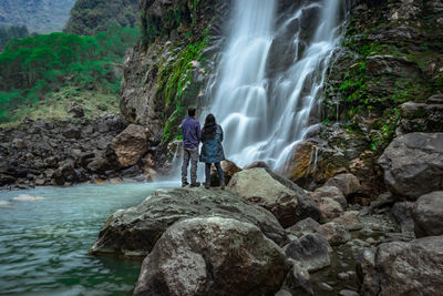 Couple standing at rock in front of waterfall white water stream falling from mountains