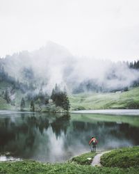 Rear view of man standing by lake in forest during foggy weather
