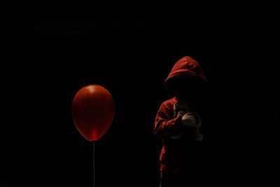 Boy wearing hood holding stuffed toy while standing by red balloon against black background