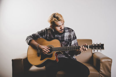 Young man playing guitar while sitting on armchair against white background