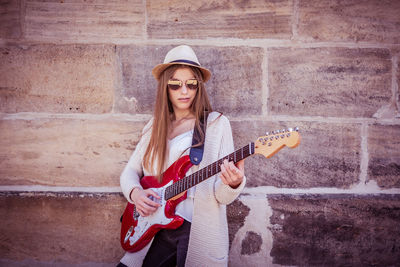 Portrait of young woman playing guitar while standing by wall