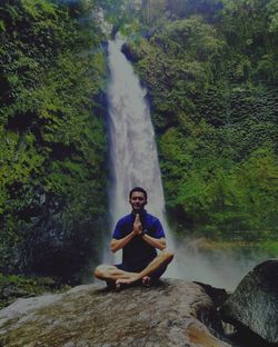 Man practicing yoga while sitting on rock against waterfall at forest