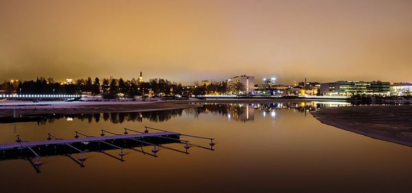 Illuminated city buildings by river against sky at sunset