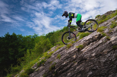 Downhill rider cyclist rides down a steep cliff surrounded by green trees against a blue sky. copy