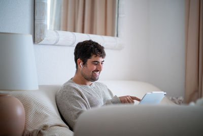 Man using digital tablet sitting on bed against wall at home
