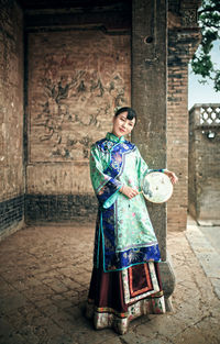 Smiling young woman with hand fan standing at old building