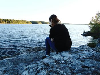 Sad woman sitting on rock at lakeshore against clear sky