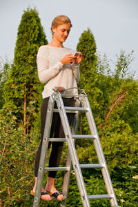 Young woman using smart phone while standing on ladder at yard