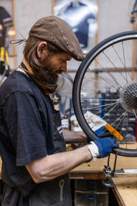 Side view of mature bearded mechanic in workwear and gloves checking and repairing wheel of bike during maintenance service in workshop